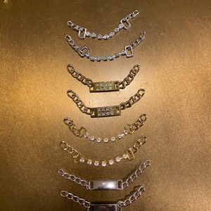 Könnte beinhalten: Neun silberne und goldene Gliederarmbänder mit verschiedenen Anhängern, darunter quadratische und rechteckige Platten mit Strasssteinen und schlichte rechteckige Platten.