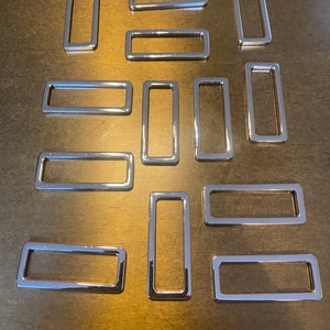 Puede incluir: Una colección de anillos metálicos rectangulares plateados y brillantes. Los anillos están dispuestos sobre una superficie de tono neutro. El metal tiene una calidad reflectante, lo que sugiere un acabado pulido. Estos anillos podrían usarse para varios proyectos de artesanía o ferretería.