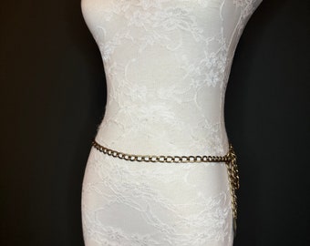 May include: A gold chain belt with a decorative pendant. The belt is on a white lace-covered mannequin.