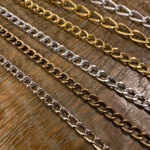 May include: Five different chain links in various metal finishes, including silver, gold, and brass. The chains are arranged in a staggered pattern on a wooden surface.