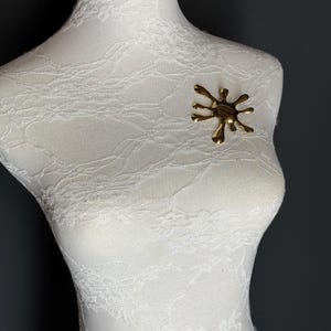 Puede incluir: Un broche de color dorado en forma de salpicadura o estrella. El broche est&aacute; en un maniqu&iacute; blanco.