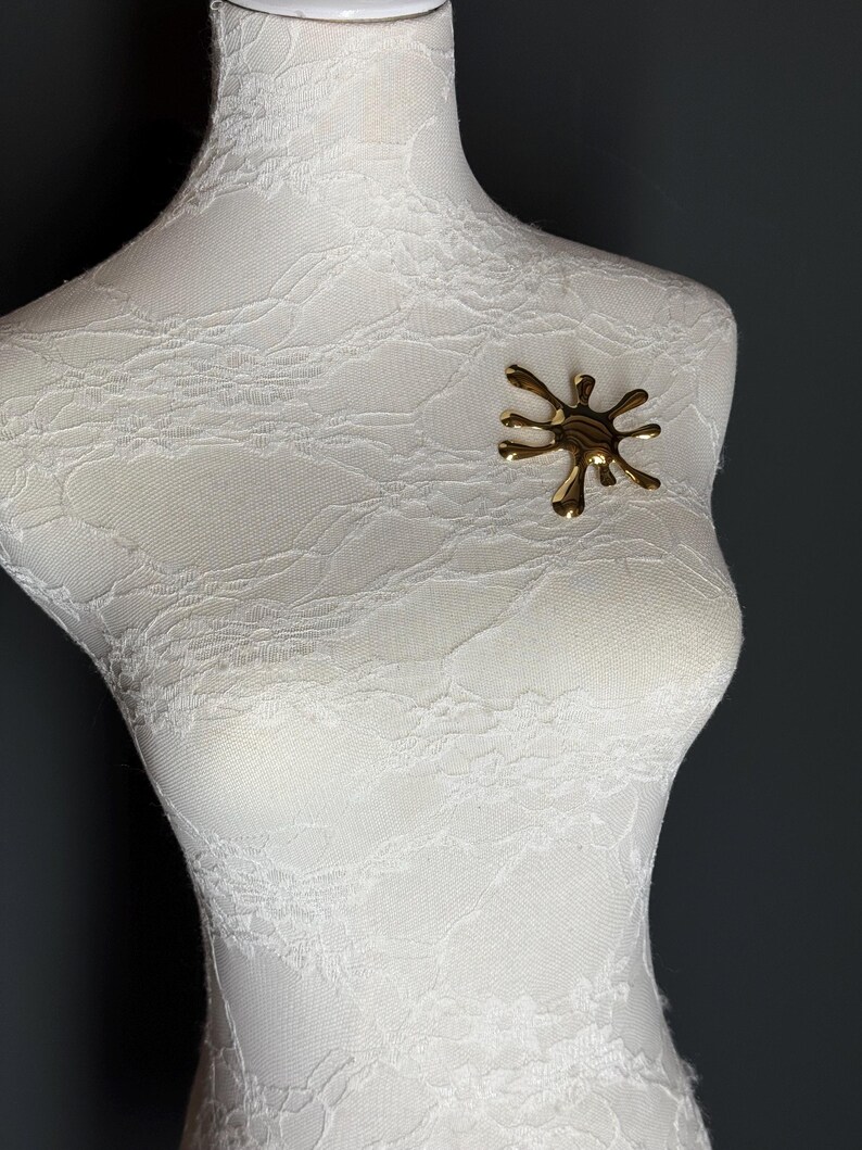 Puede incluir: Un broche de color dorado en forma de salpicadura o estrella. El broche est&aacute; en un maniqu&iacute; blanco.