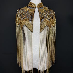 May include: A gold beaded cape with a high collar and intricate floral embroidery. The cape features long, dangling beaded fringe. The garment is displayed on a white mannequin against a black background, showcasing its elegant design.