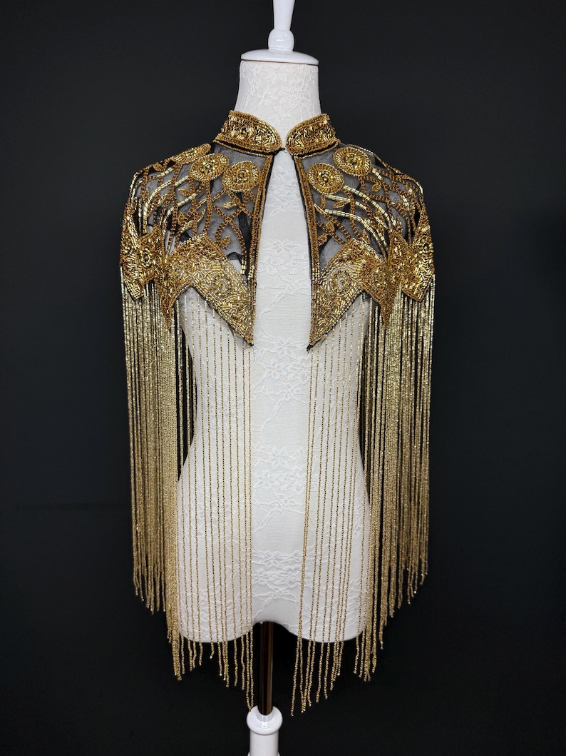 May include: A gold beaded cape with a high collar and intricate floral embroidery. The cape features long, dangling beaded fringe. The garment is displayed on a white mannequin against a black background, showcasing its elegant design.