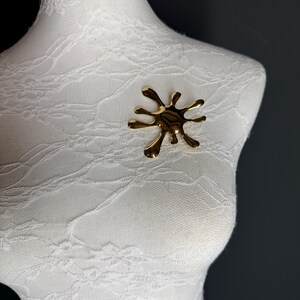 Puede incluir: Un broche abstracto de color dorado en forma de salpicadura o gota. El broche est&aacute; en un maniqu&iacute; blanco.