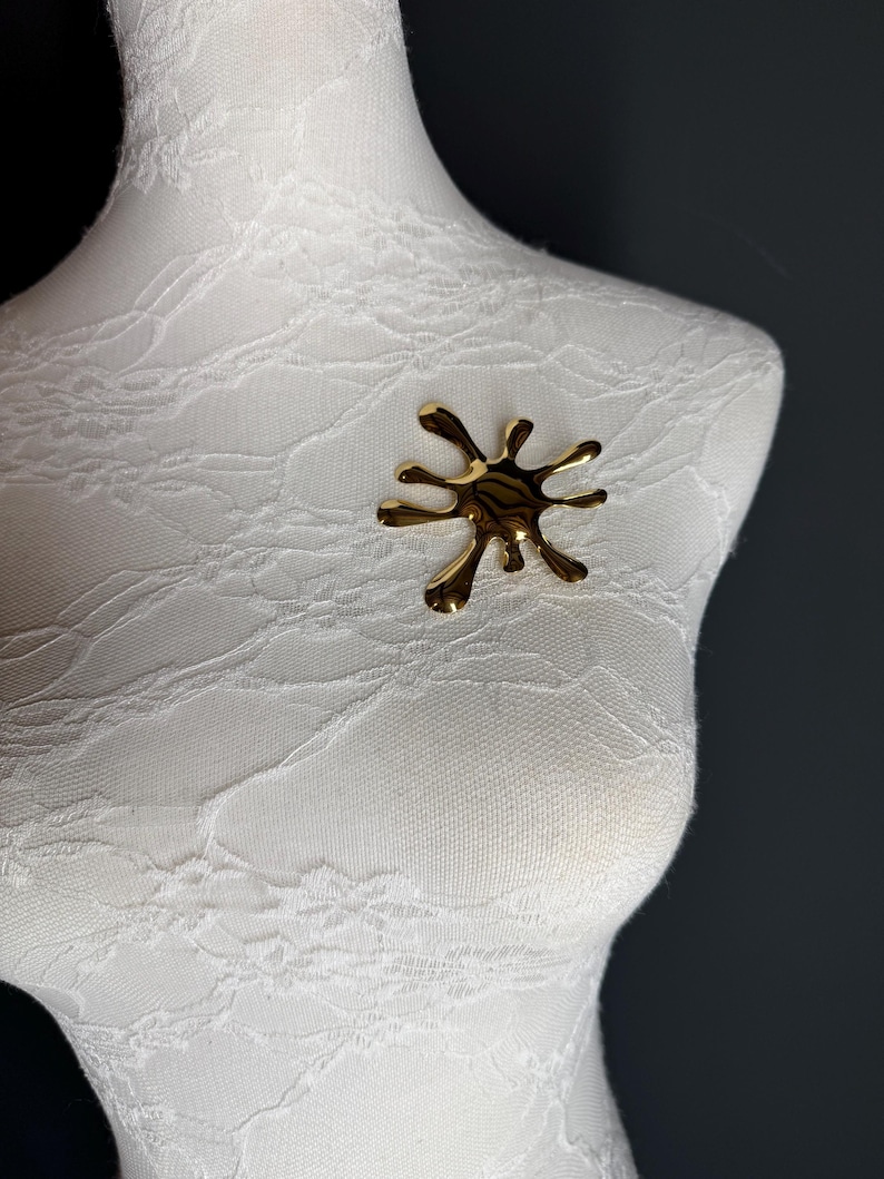 Puede incluir: Un broche abstracto de color dorado en forma de salpicadura o gota. El broche est&aacute; en un maniqu&iacute; blanco.