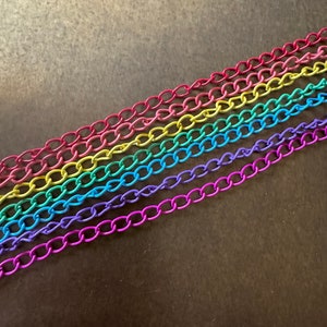 Puede incluir: Primer plano de seis eslabones de cadena coloridos en un arcoíris de colores: rojo, amarillo, verde, azul y morado. Las cadenas están dispuestas diagonalmente sobre una superficie marrón oscuro. Las cadenas son de metal.