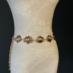 May include: A gold chain belt with a series of scallop-shaped metal charms. The belt is on a white mannequin.