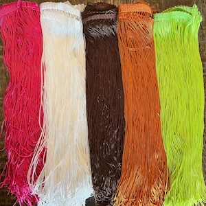 Op de afbeelding: Vijf verticale stroken franje die naast elkaar in een rij zijn geplaatst. De kleuren van links naar rechts zijn felroze, wit, donkerbruin, koperoranje en neon limoengroen. Elke strook heeft een geweven bovenrand met lange draden.