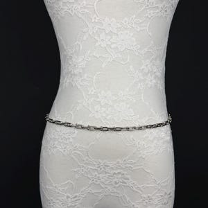 Op de afbeelding: Een zilverkleurige tailleketting met een reeks uniforme, rechthoekige schakels verbonden door kleine, afgeronde bolletjes. De ketting wordt horizontaal weergegeven over het midden van een witte, met kant bedekte etalagepop tegen een zwarte achtergrond.
