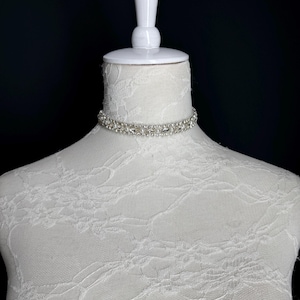 May include: A silver choker necklace with clear rhinestones is displayed on a white mannequin. The necklace is made of multiple rows of small, sparkling stones. The mannequin is covered in white lace fabric, and the background is black.