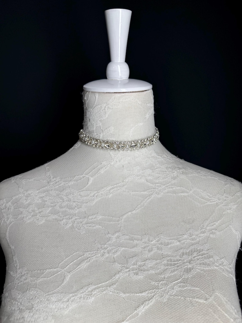 May include: A silver choker necklace with clear rhinestones is displayed on a white mannequin. The necklace is made of multiple rows of small, sparkling stones. The mannequin is covered in white lace fabric, and the background is black.