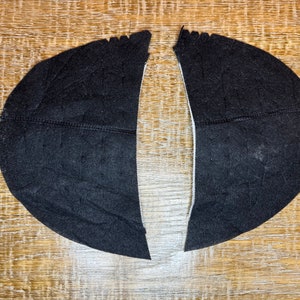 Op de afbeelding: Twee zwarte stofstukken, in een halve cirkelvorm gesneden, met een rechte rand aan één kant. De stofstukken liggen op een houten oppervlak.