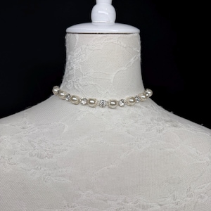 Puede incluir: Un delicado collar gargantilla con perlas de imitación y cristales transparentes. El collar se exhibe en un maniquí blanco sobre un fondo negro, resaltando el elegante diseño de la joya.