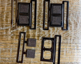 May include: Four metal belt buckles with a rectangular design. Each buckle has a dark gray mesh center and a silver-colored frame. The buckles are arranged on a wooden surface.
