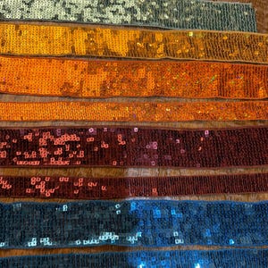 Puede incluir: Una colección de cintas con adornos de lentejuelas en varios colores, incluyendo plata, oro, naranja, rojo y azul. Las lentejuelas están densamente empaquetadas, creando un efecto brillante. Las cintas están dispuestas horizontalmente, mostrando la gama de colores y texturas.