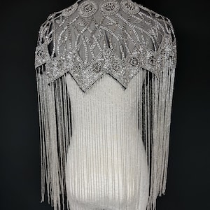 May include: A silver beaded cape with a high collar and detailed shoulder embellishments. The cape has a sheer, net-like fabric decorated with circular and geometric designs. Long beaded strands hang down, forming a fringe. The garment is shown on a white mannequin against a black background.
