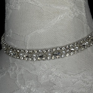 May include: A sparkling choker necklace with clear crystals and small white pearls. The necklace is set on a white fabric form, showcasing its design. The crystals are arranged in a row, adding a touch of elegance.