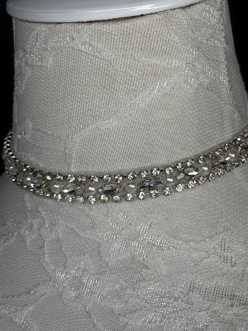 May include: A sparkling choker necklace with clear crystals and small white pearls. The necklace is set on a white fabric form, showcasing its design. The crystals are arranged in a row, adding a touch of elegance.