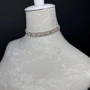 May include: A sparkling, double-strand rhinestone choker necklace. The necklace is displayed on a white mannequin head with a lace pattern. The background is a dark, solid color.