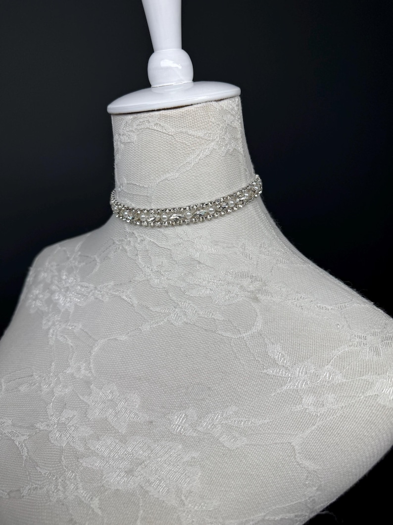 May include: A sparkling, double-strand rhinestone choker necklace. The necklace is displayed on a white mannequin head with a lace pattern. The background is a dark, solid color.