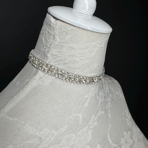 May include: A silver choker necklace with clear crystals and small white pearls. The necklace is displayed on a white mannequin head with a lace fabric covering. The background is black.