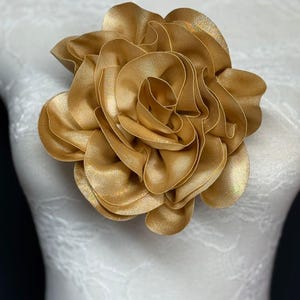 Puede incluir: Una gran flor de tela de color amarillo dorado con múltiples capas con volantes. La flor está unida a un maniquí blanco. Los pétalos tienen un brillo sutil, creando una apariencia lujosa. Este accesorio floral podría usarse como broche o adorno para el cabello.