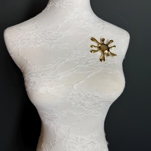 Puede incluir: Un broche abstracto de color dorado en forma de salpicadura o salpicadura. El broche est&aacute; prendido a un maniqu&iacute; cubierto de encaje blanco.