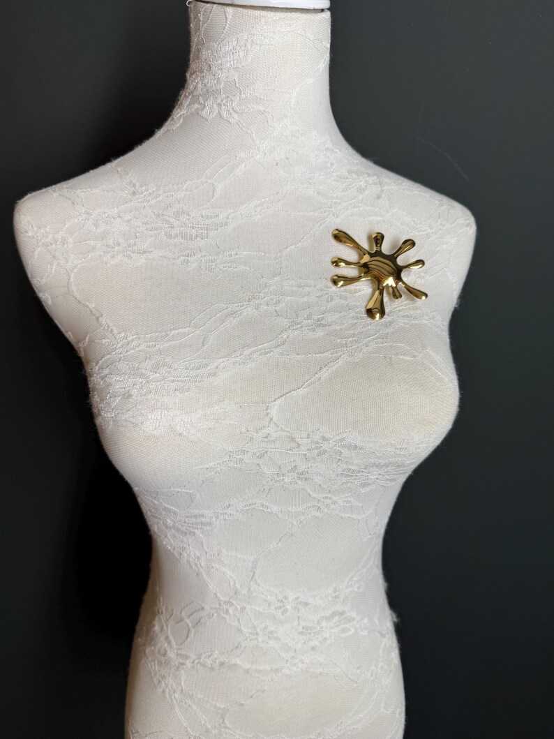 Puede incluir: Un broche abstracto de color dorado en forma de salpicadura o salpicadura. El broche est&aacute; prendido a un maniqu&iacute; cubierto de encaje blanco.