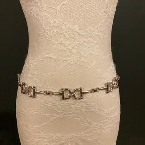 May include: A silver chain belt with a repeating pattern of interlocking oval links. The belt is displayed on a white lace-covered mannequin.
