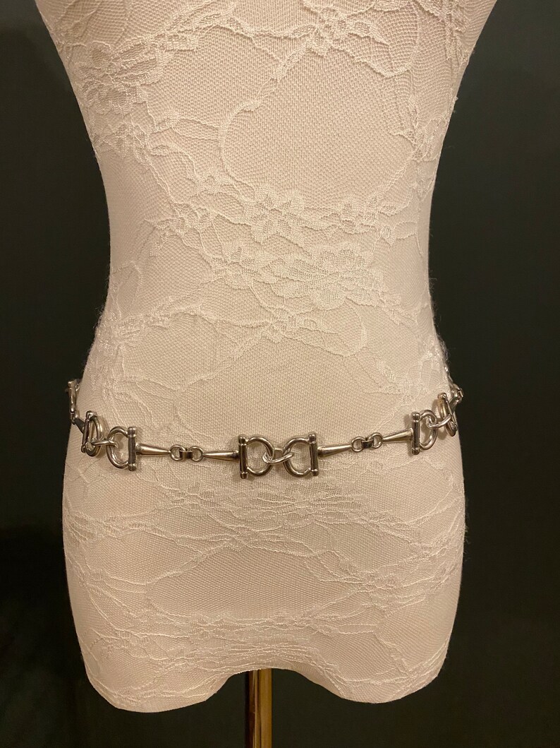 May include: A silver chain belt with a repeating pattern of interlocking oval links. The belt is displayed on a white lace-covered mannequin.