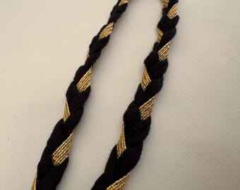 May include: Close-up of two black braided cords with gold metallic accents. The cords are intertwined, creating a textured pattern. The gold accents add a touch of elegance to the design.