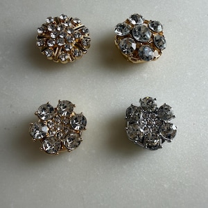 May include: Four decorative buttons with a floral design. Two buttons have a gold-tone base, and two have a silver-tone base. Each button is embellished with clear, sparkling rhinestones, creating a glamorous look.