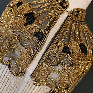 May include: Close-up of a gold beaded collar with black accents. The collar is made of a sheer black fabric and is heavily embellished with gold beads in intricate patterns. Gold fringe hangs from the bottom edge. The collar is displayed on a white form.