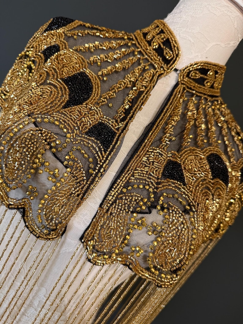 May include: Close-up of a gold beaded collar with black accents. The collar is made of a sheer black fabric and is heavily embellished with gold beads in intricate patterns. Gold fringe hangs from the bottom edge. The collar is displayed on a white form.