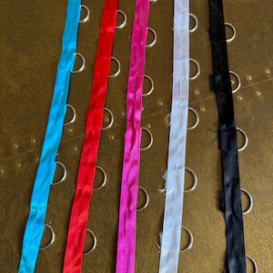 Puede incluir: Una colección de cremalleras coloridas en tonos azul, rojo, rosa, blanco y negro. Las cremalleras están dispuestas verticalmente sobre una superficie dorada texturizada, con anillos metálicos visibles.