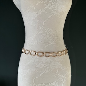 May include: A gold chain belt with oval links. The belt is on a white lace-covered mannequin.