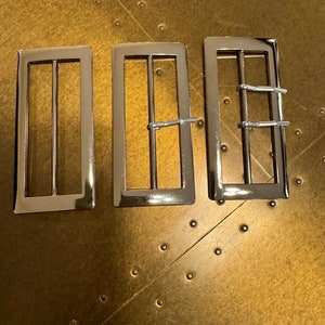 Puede incluir: Tres hebillas de cinturón de metal plateado con diferentes diseños. Las hebillas son rectangulares y tienen un acabado pulido.