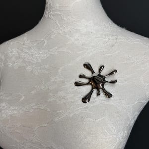 Puede incluir: Un broche plateado en forma de una salpicadura abstracta estilizada. El broche est&aacute; hecho de metal y tiene un acabado brillante.
