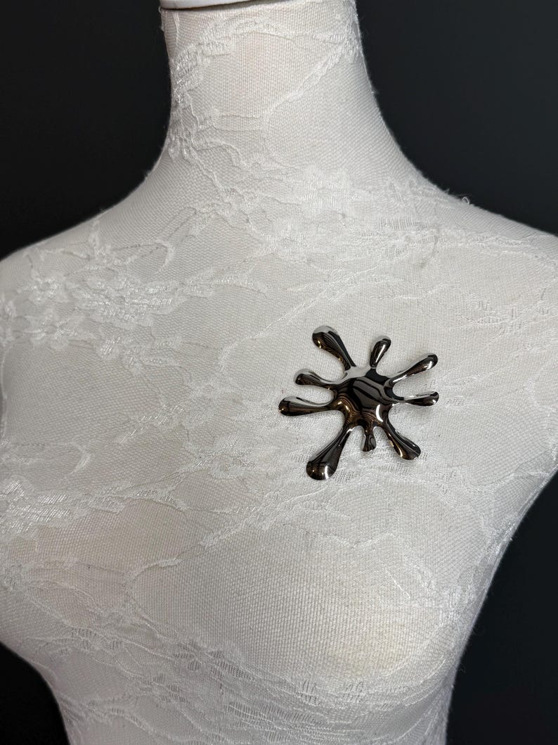 Puede incluir: Un broche plateado en forma de una salpicadura abstracta estilizada. El broche est&aacute; hecho de metal y tiene un acabado brillante.