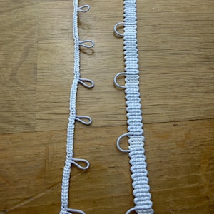 May include: Two strips of white curtain heading tape. The left strip is a narrow woven band with small, evenly spaced loops extending from one side. The right strip is a wider, thicker woven band featuring larger, semi-circular loops along one edge.