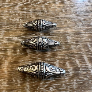 Könnte beinhalten: Drei silberne Metallperlen mit einem verzierten Design. Die Perlen sind oval geformt und haben ein erhabenes Muster.