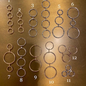 May include: Twelve different styles of gold and silver chain links. Each chain link is made of metal and has a circular shape. The chain links are arranged in rows on a brown surface.