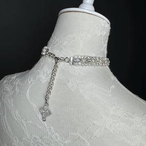 May include: A silver choker necklace with a rhinestone and pearl design. The necklace is on a white mannequin. The choker has a chain extender and a rhinestone pendant. The necklace is a delicate piece of jewelry.