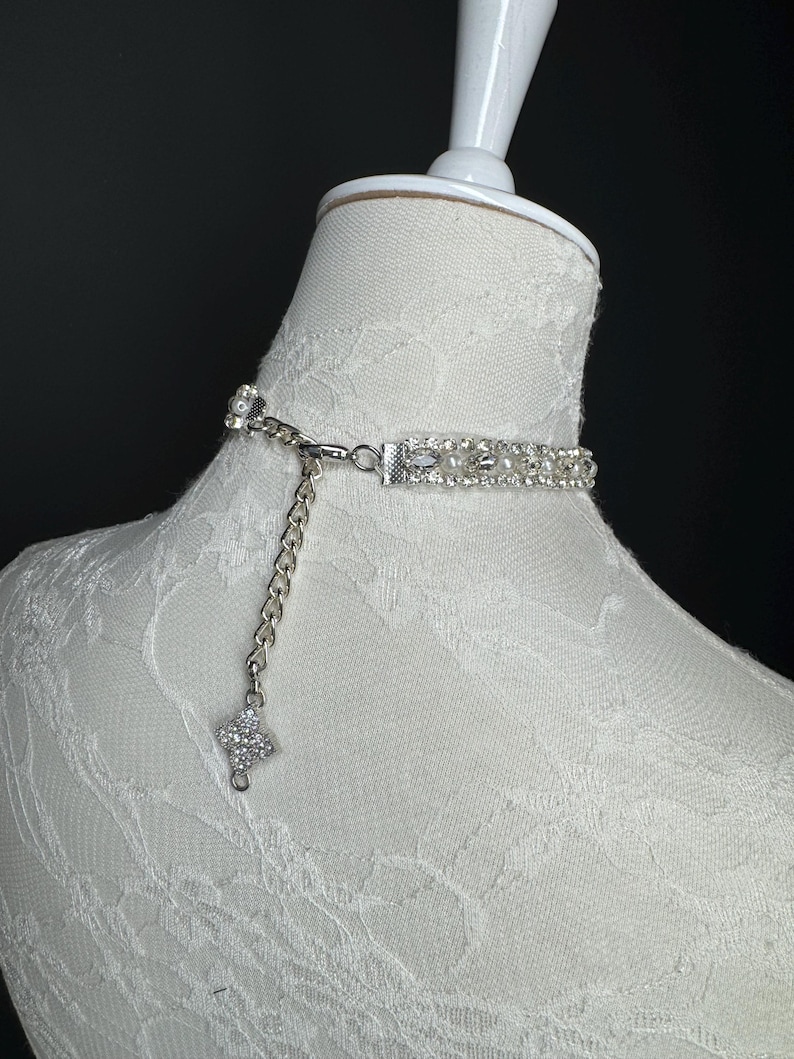 May include: A silver choker necklace with a rhinestone and pearl design. The necklace is on a white mannequin. The choker has a chain extender and a rhinestone pendant. The necklace is a delicate piece of jewelry.
