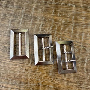 Könnte beinhalten: Drei silberfarbene rechteckige Gürtelschnallen mit klassischem Design. Jede Schnalle hat einen Rahmen mit einem einzelnen oder doppelten Steg für den Gürtelriemen. Die Schnallen werden auf einer Holzoberfläche präsentiert.