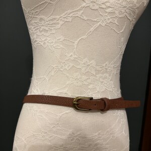 Op de afbeelding: Een bruine leren riem met een gouden gesp. De riem is op een witte kanten mannequin.