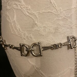 May include: A silver chain belt with multiple interlocking oval-shaped buckles. The belt is on a white fabric background.