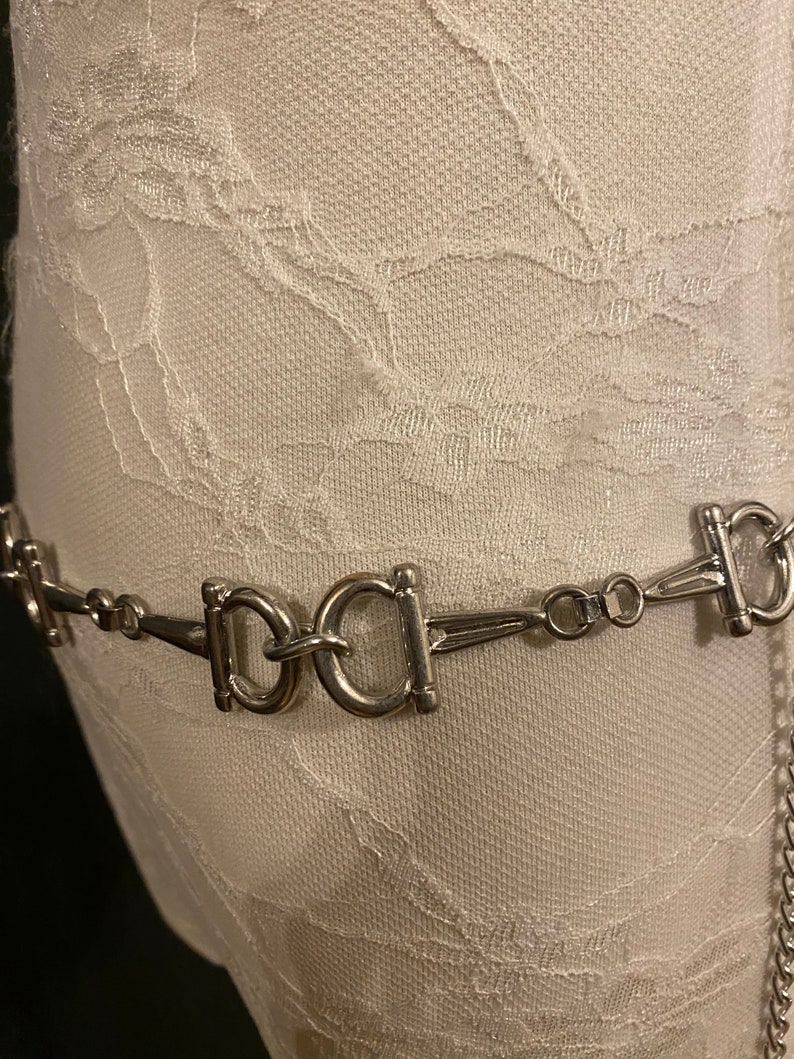 May include: A silver chain belt with multiple interlocking oval-shaped buckles. The belt is on a white fabric background.