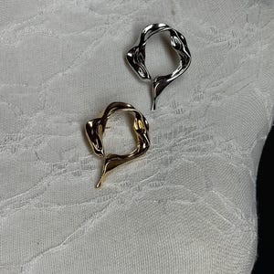 May include: Two abstract metal rings, one gold and one silver, with a wavy, organic design. The rings are displayed on a white fabric background.